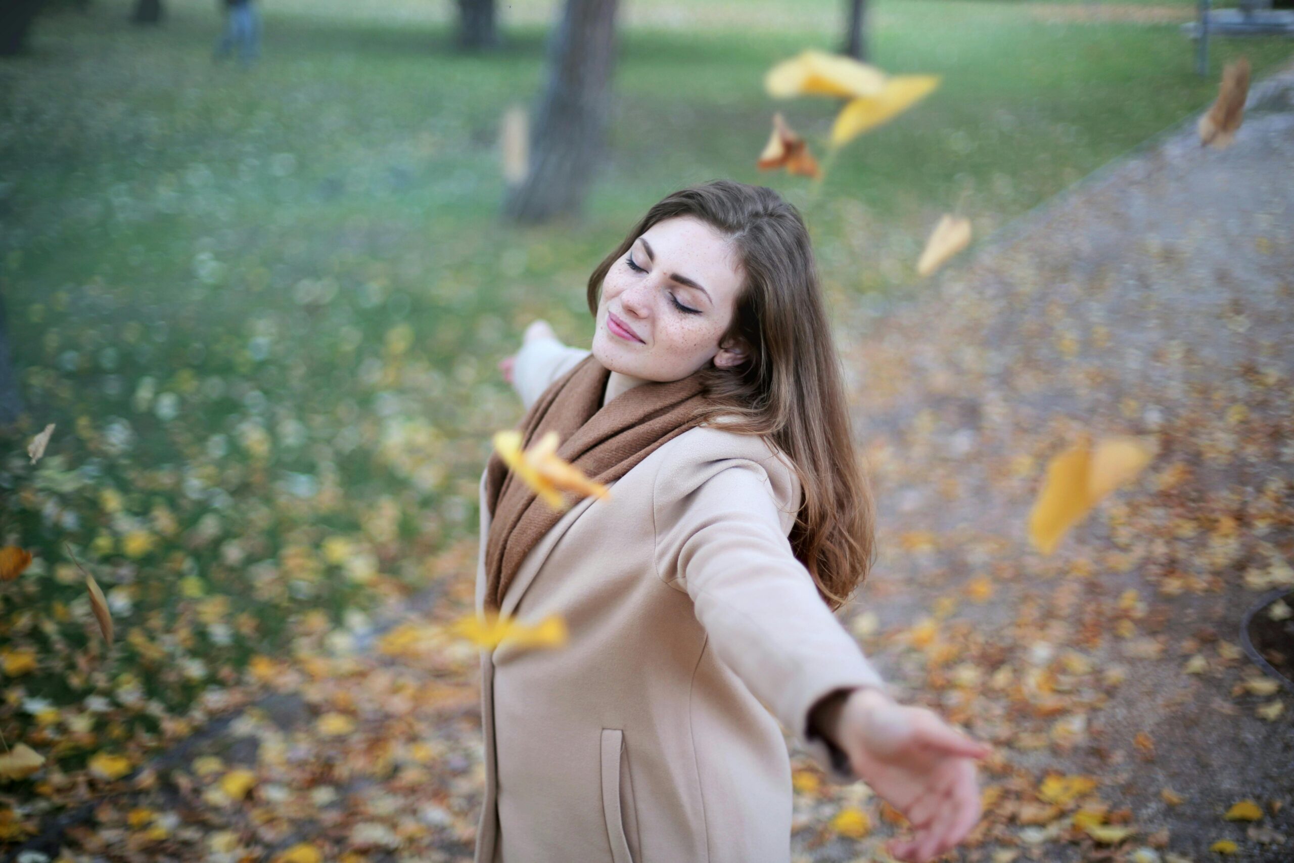 Happy woman with arms outstretched enjoying autumn leaves falling in a park, embracing the season with joy.