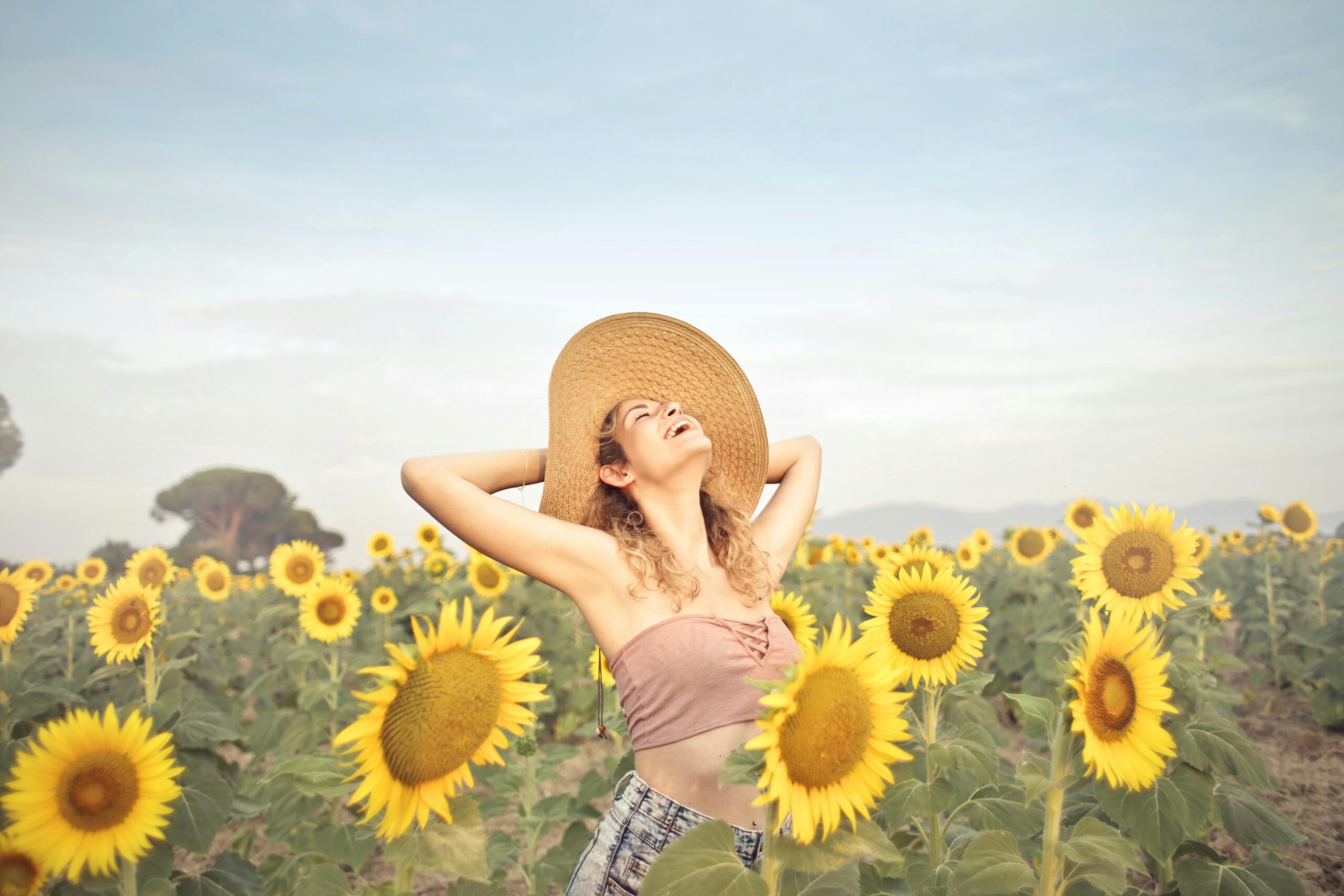 Smiling woman in a sunflower field wearing a wide-brimmed hat and pink top, enjoying the sunshine and nature.